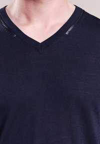 Navyblau gestrickter V-Ausschnitt-Pullover mit geripptem Kragen und subtiler glanzender Oberfläche. Verfügt über markante Schulterakzente aus einem kontrastierenden Material.