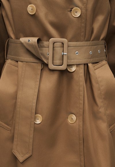Καφέ παλτό τύπου τραντσκότ με ζώνη από το ίδιο ύφασμα, ορθογώνια αγκράφα, διπλή σειρά κουμπιών και πλαϊνές τσέπες.
