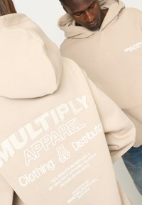 Felpa beige con ricamo del testo "MULTIPLY APPAREL" e logo. Tessuto morbido con cappuccio strutturato e vestibilità ampia.