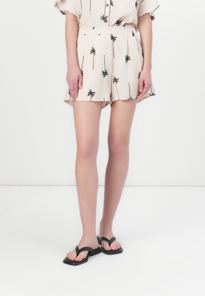 Shorts beiges avec des imprimés de palmiers noirs, présentant une coupe décontractée, une taille élastique et un design plissé. Associés à des tongs noires.