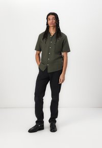 Camisa de manga corta, verde oscuro, abotonada, con un tejido texturizado. Combinada con pantalones negros y zapatos negros, de pie contra un fondo blanco.