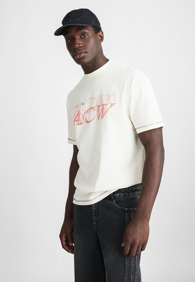 Camiseta de manga corta crema con el texto gráfico rojo "ACW" en el frente, combinada con jeans de denim negros que tienen detalles de costura lateral.
