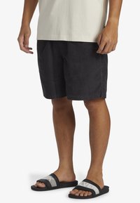 Sorte kordfløjs shorts med sidelommer, iført sorte slidesandaler med en stribet grå bånd over toppen.