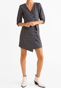 Robe portefeuille à carreaux gris avec manches trois-quarts, dotée d'un col en V, de détails à boutons et d'un ourlet asymétrique. Associée à des bottines blanches.