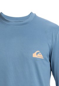 Modrá košeľa s dlhým rukávom z hladkej tkaniny. Má jemný okrúhly výstrih a svetlooranžové logo na hornej ľavej časti hrude.