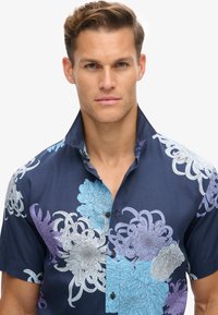 Camisa de manga corta en tejido azul marino con grandes patrones florales en azul claro y púrpura. Cierre frontal de botones con cuello de punta.