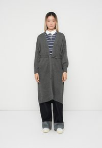 Grau gestrickte Strickjacke mit strukturiertem Muster, offenem Vorderteil und Bindegürtel. Gestylt über einem gestreiften Hemd und kombiniert mit dunkelblauen weit geschnittenen Hosen und weißen Schuhen.