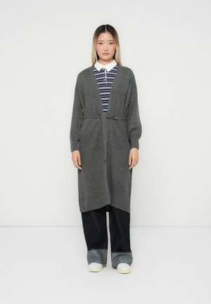 Cardigan en tricot gris avec un motif texturé, à ouverture devant et ceinture à nouer. Stylé sur une chemise rayée et associé à un pantalon large noir et des chaussures blanches.
