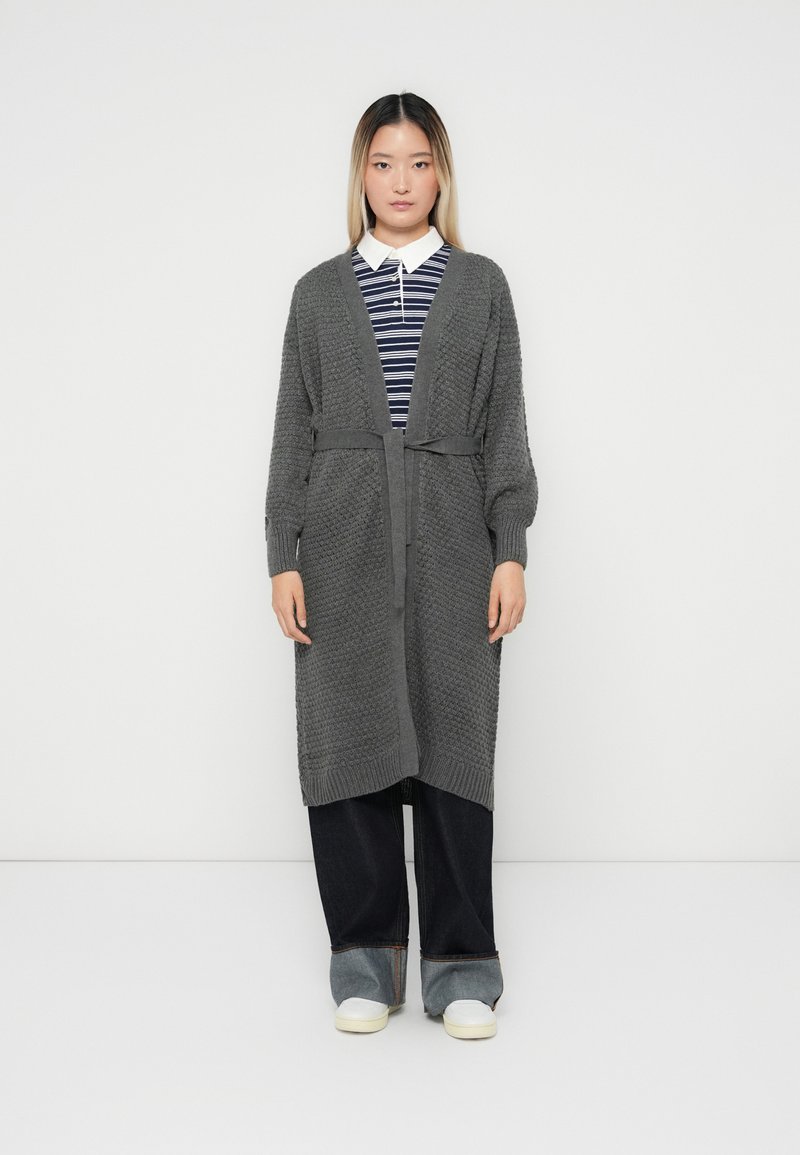 Cardigan en tricot gris avec un motif texturé, à ouverture devant et ceinture à nouer. Stylé sur une chemise rayée et associé à un pantalon large noir et des chaussures blanches.