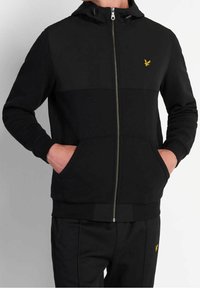 Man in een zwarte zip-up hoodie en bijpassende zwarte broek, met zijn handen in de zakken, met kleine gele adelaar logo's op de borst en broek.