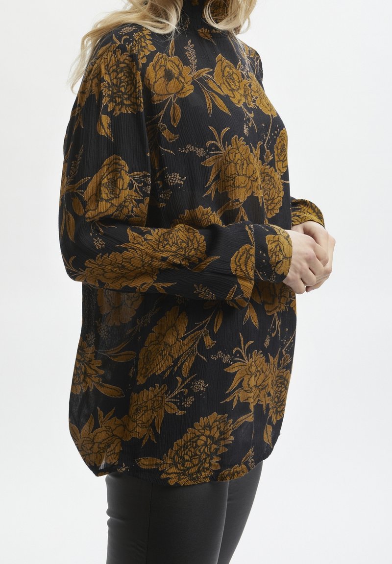 Femme portant un chemisier noir à manches longues avec des motifs floraux jaune moutarde, associée à un pantalon noir, debout devant un fond blanc.