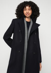 Palton bleumarin cu două rânduri de nasturi, cu o croială ajustată, rever în V și nasturi negri. Purtați peste o eșarfă gri pentru un contrast suplimentar.