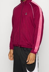 Burgundy zipjacka med rosa sidestreck, utrustad med ståkrage, elastiska ärmslut och logotypdetaljer på bröstet. Tillverkad av mjukt tyg.