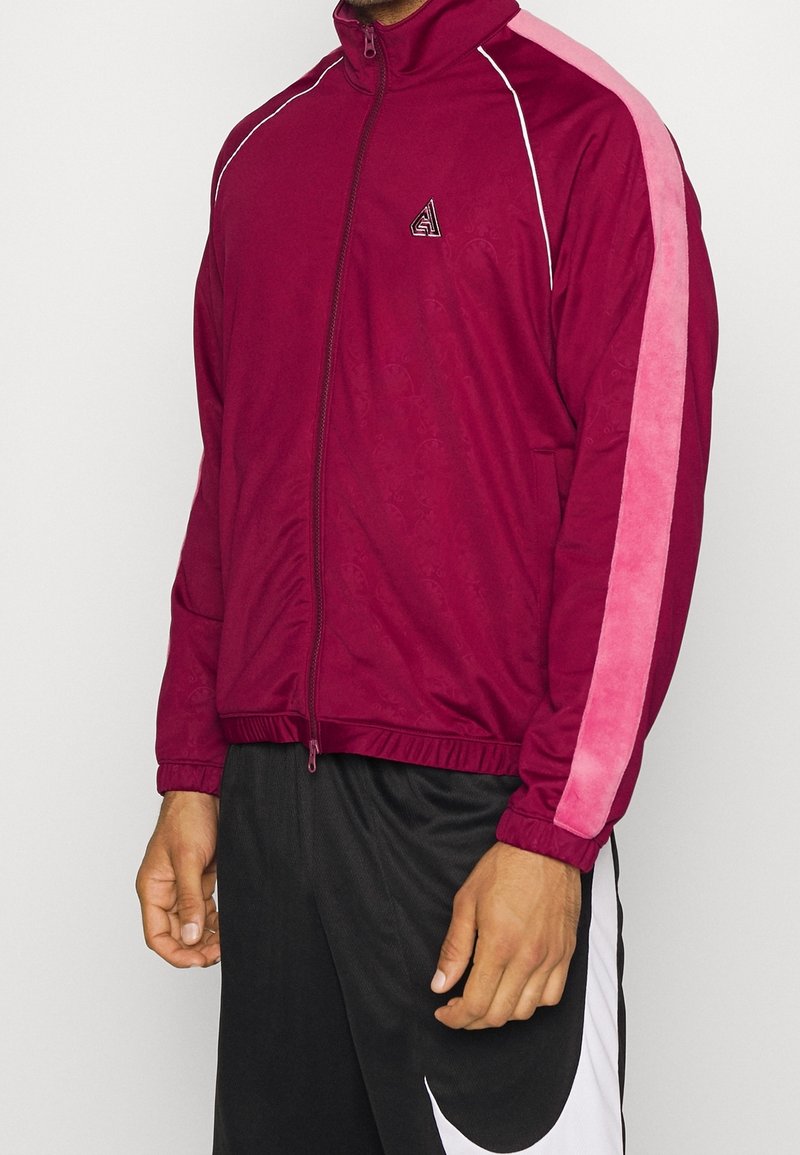 Burgundy zipjacka med rosa sidestreck, utrustad med ståkrage, elastiska ärmslut och logotypdetaljer på bröstet. Tillverkad av mjukt tyg.