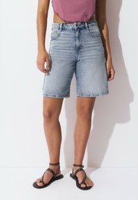 Shorts de mezclilla azul claro con un corte relajado, dobladillo desgastado y bolsillos frontales; combinados con sandalias planas negras con acentos de tachuelas.