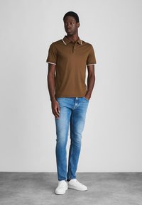 Polo shirt maron cu guler și accente albe la mâneci, asortat cu blugi ușor albaștri și teniși albi. Modelul stă pe un podea gri.