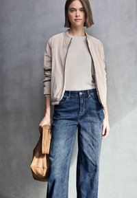 Chaqueta bomber de color beige claro con cremallera frontal, combinada con una blusa ceñida de color crema y jeans de pierna ancha azules. Sosteniendo un bolso de cuero marrón.