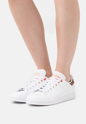 Białe skórzane sneakersy z perforowanymi akcentami, kontrastującymi pomarańczowymi i czerwonymi oczkami oraz wzorzystą pętelką na pięcie. Okrągły czubek i płaska podeszwa.