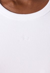 Haut en tissu côtelé blanc avec un col rond. Présente un logo brodé discret en blanc. Texture lisse avec des lignes verticales.