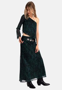 Vestido asimétrico de un solo hombro en verde oscuro con una tela fluida y transparente. Presenta un dobladillo asimétrico y un cinturón plateado con acentos circulares. Botines negros hasta el tobillo.
