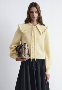 Gelbes Strickpullover mit einem Rüschenausschnitt, Knopfverschluss und langen Ärmeln, kombiniert mit einem schwarzen Rock und einer braunen Handtasche im Schlangenmuster.