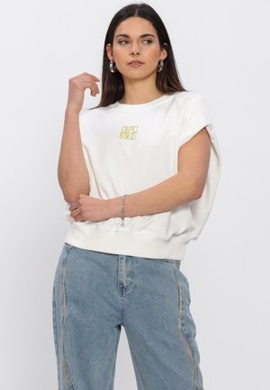 Vrouw met een wit mouwloos shirt met een geel logo en blauwe spijkerbroek met decoratieve naden, poseert met gekruiste armen en kijkt naar rechts.