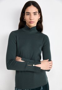 Dark green long-sleeve top with a high collar, featuring cut-out accents at the cuffs and a smooth, lightweight texture.