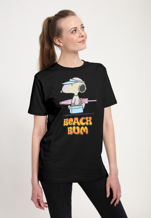 Jonge vrouw die een zwart T-shirt draagt met Snoopy die een strandparasol en koelbox vasthoudt, tekst "BEACH BUM" in oranje en gele letters.