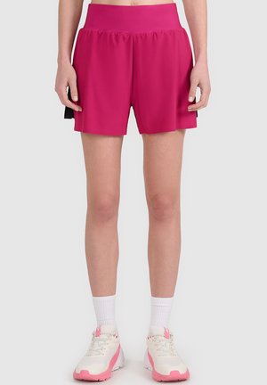 Mujer vistiendo pantalones cortos deportivos rosa brillante, calcetines blancos y zapatillas blancas con detalles rosas sobre un fondo gris claro.