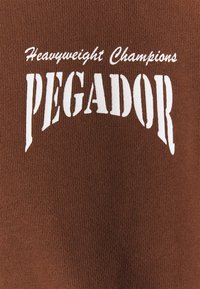 Hnědá bavlněná tkanina s výrazným bílým textem: "Těžká váha šampióni PEGADOR." Text má zakřivený design. Hladká textura.