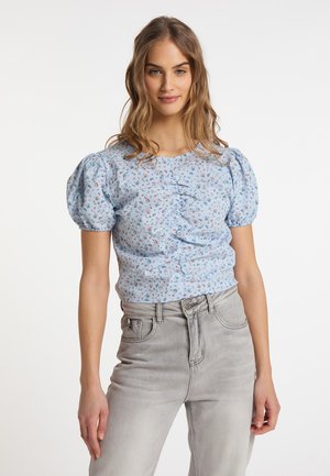 Junge Frau trägt eine hellblaue, geblümte Bluse mit Puffärmeln und hellgraue, hoch taillierte Jeans, steht vor einem schlichten Hintergrund.