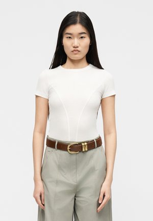 Kobieta z długimi ciemnymi włosami, ubrana w dopasowaną białą bluzkę z krótkim rękawem, jasnoszare spodnie i brązowy skórzany pasek z złotą klamrą.