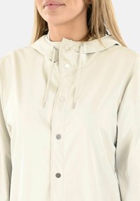 Chaqueta impermeable de color beige claro con capucha, cierre de botones a presión, cordones y una textura suave y mate. Presenta un diseño minimalista.