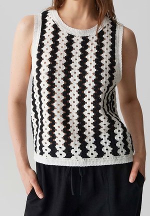 Femme portant un haut sans manches en crochet blanc avec des rayures verticales noires et un pantalon noir, mains dans les poches, debout devant un fond uni.