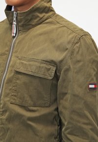 Olive green bomber jacket with a high collar, front zipper, and a chest pocket; features a logo patch on the left sleeve.