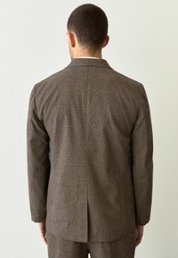 Braun kariertes Blazer mit taillierter Passform, ausgestattet mit einem Schlitz auf der Rückseite und passenden Hosen, die unten sichtbar sind.