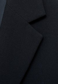 Črn teksturiran material, ki prikazuje ostre, angularne robove klap in prilagojene detajle, kar poudarja strukturiran dizajn oblačila.