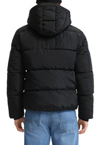 Plumífero negro con capucha, con acolchado horizontal y puños elásticos. Llevado sobre jeans de mezclilla azul.