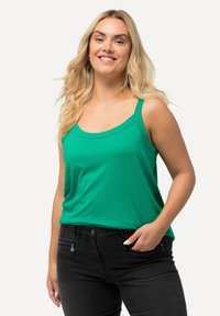 Top senza maniche verde realizzato in un tessuto morbido con scollo rotondo e vestibilità comoda, abbinato a pantaloni neri con tasche con zip.