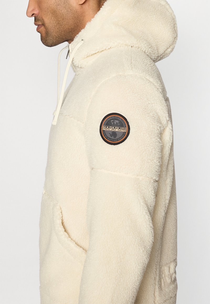 Mann in einer cremefarbenen Fleece-Hoodie mit Fronttasche und kreisförmigem Napapijri-Logopatch am Oberarm, der nach links in einer Nahaufnahme seitlich blickt.