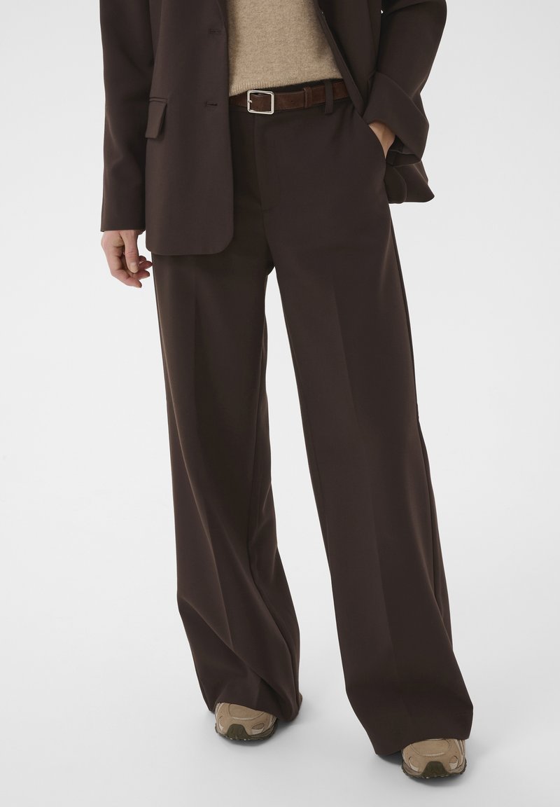 Personne portant un costume ample marron foncé, un pull beige, une ceinture marron et des baskets beiges, avec une main dans la poche sur un fond uni.