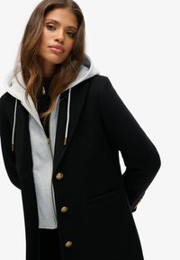 Zwarte wollen coat met gouden knopen, een ingeklapte kraag en een grijze hoodie eronder. Zachte textuur met een getailleerde pasvorm.