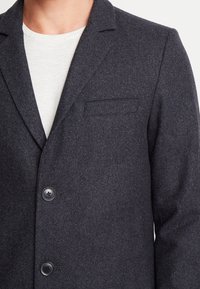 Uomo che indossa un blazer di lana grigio scuro con due bottoni neri sopra una maglietta bianca a collo rotondo, torso visibile, sfondo neutro.