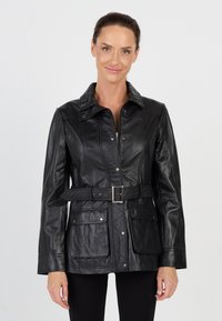 Kobieta w czarnej skórzanej kurtce z paskiem i kieszeniami na klapie, stojąca na jednolitym białym tle, w czarnych spodniach.