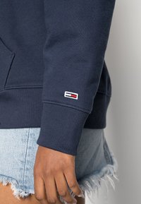Sweatshirt bleu marine avec des poignets côtelés, arborant une petite patch logo multicolore sur la manche gauche. Associé à un short en denim effiloché.