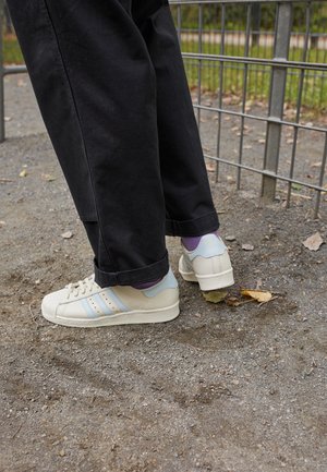 Personne portant des baskets blanches avec des rayures bleu clair et des chaussettes violettes, debout sur un chemin de gravier près d'une clôture métallique.