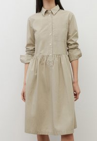 Abito camicia in cotone beige con colletto a bottone, maniche lunghe e vita arricciata. Presenta una tasca sul petto e un tessuto testurizzato.