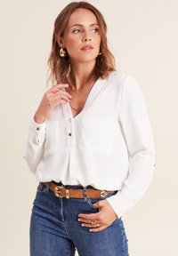 Blouse blanche à manches longues avec un décolleté en V, deux poches poitrine et des boutons décoratifs. Associée à un jean bleu et une ceinture marron.