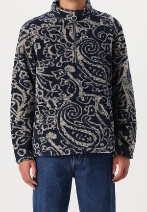 Granatowo-beżowy polarowy sweter z nadrukiem, z półzamkiem i stojącym kołnierzem, noszony z niebieskimi dżinsami.