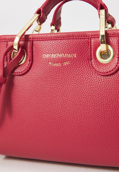 Bolso de mano de cuero rojo con textura de guijarros y herrajes dorados, que cuenta con asas dobles y un logotipo en relieve. Los detalles notables incluyen ojetes y un acabado texturizado.